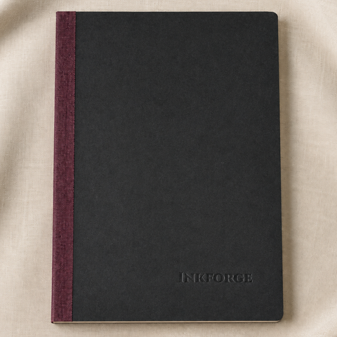 A black hardcover notebook with a linen burgundy spine, placed on a beige fabric background. The brand name 'INFORGE' is embossed on the bottom right corner of the cover.