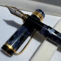 A luxury fountain pen with a glossy black and blue marbled resin body and gold-plated nib and trim, resting on a white velvet surface.