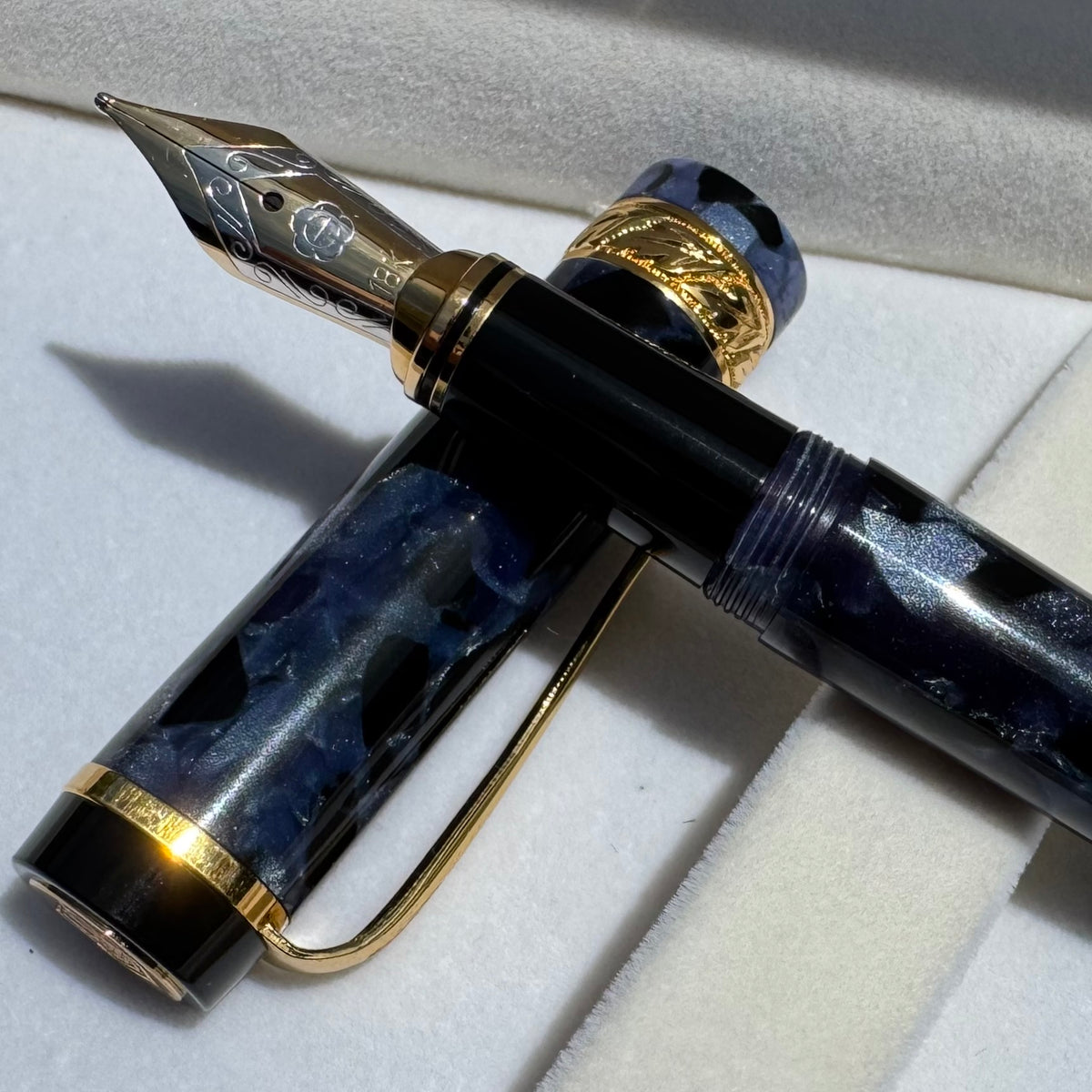 A luxury fountain pen with a glossy black and blue marbled resin body and gold-plated nib and trim, resting on a white velvet surface.