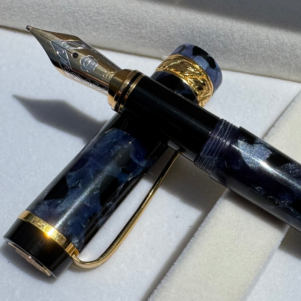 A luxury fountain pen with a glossy black and blue marbled resin body and gold-plated nib and trim, resting on a white velvet surface.