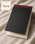 A black notebook with a burgundy top band and the brand name 'INKFORGE' debossed near the bottom, resting on a cream-colored book or folder. The scene is styled on draped beige fabric, with a cream label in the top-left corner reading 'Aura'.
