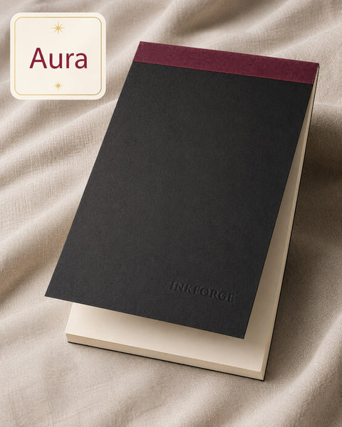 A black notebook with a burgundy top band and the brand name 'INKFORGE' debossed near the bottom, resting on a cream-colored book or folder. The scene is styled on draped beige fabric, with a cream label in the top-left corner reading 'Aura'.