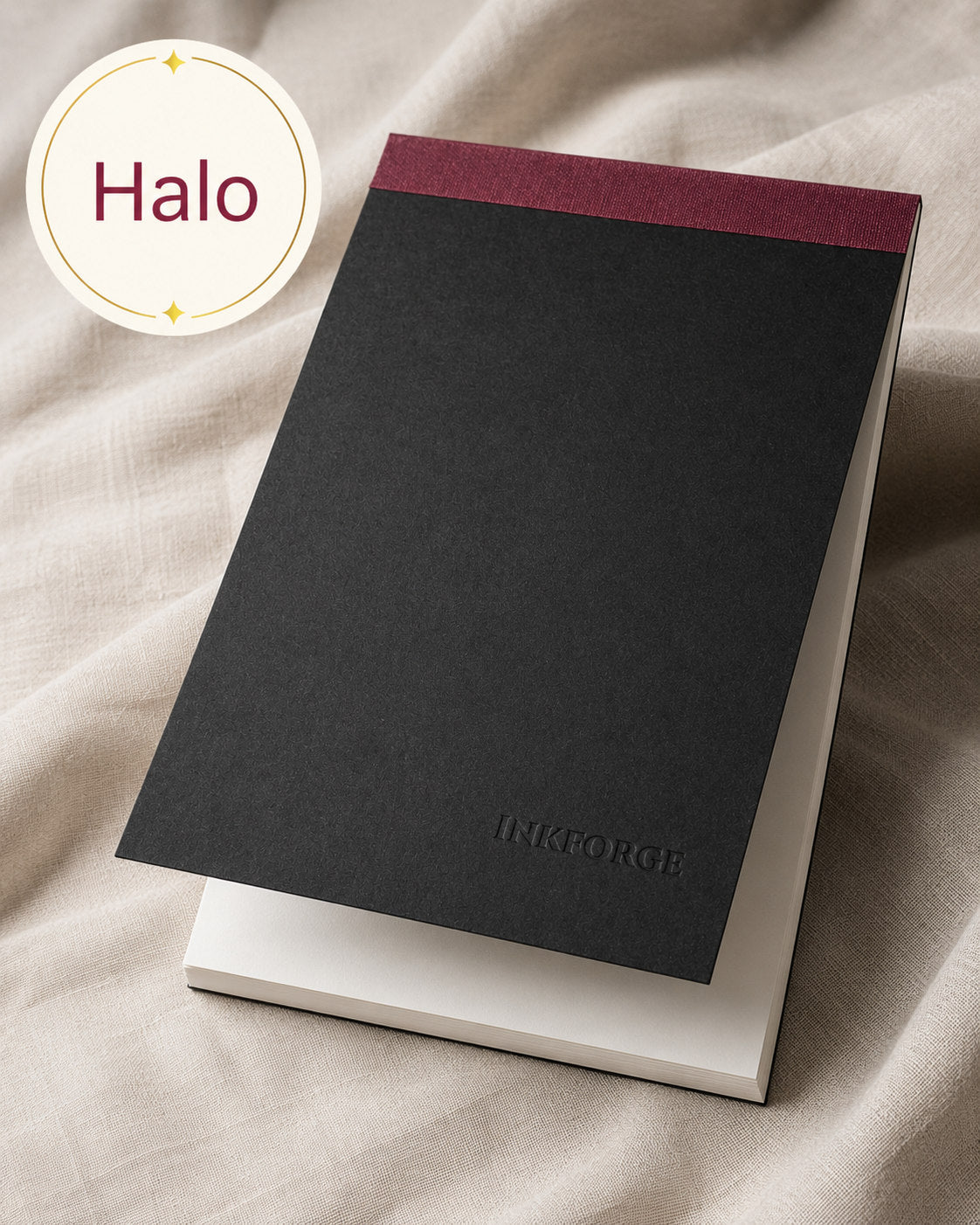 A black textured notepad with a burgundy top border and the brand name 'INKFORGE' debossed near the bottom, displayed on a white sheet over draped beige fabric. A circular badge in the top-left reads 'Halo'.